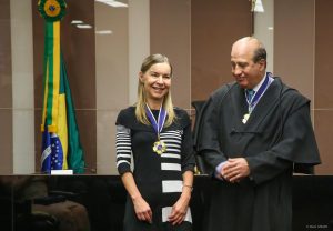 Vestindo vestido preto com listras brancas, Dra. Lúcia Willadino Braga sorri ao receber o Grande-Colar do Mérito do Tribunal de Contas da União.