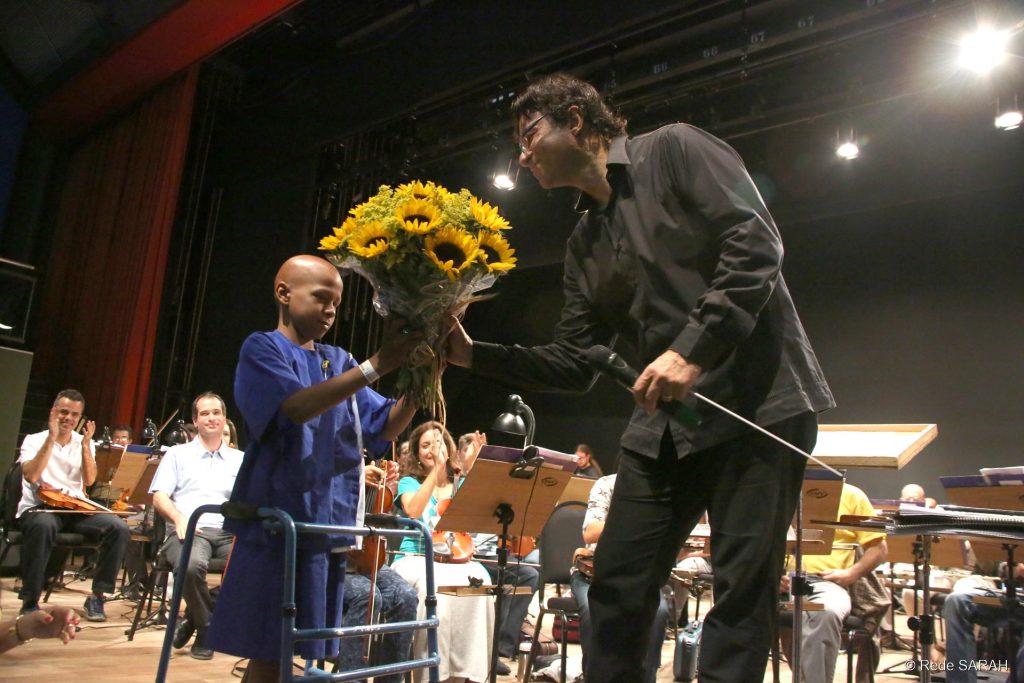 Maestro Claudio Cohen recebendo flores de um paciente do SARAH Brasília Após apresentação do espetáculo “A Música do Cinema”, o maestro Cláudio Cohen recebe um buquê de flores de menino paciente do SARAH Brasília.