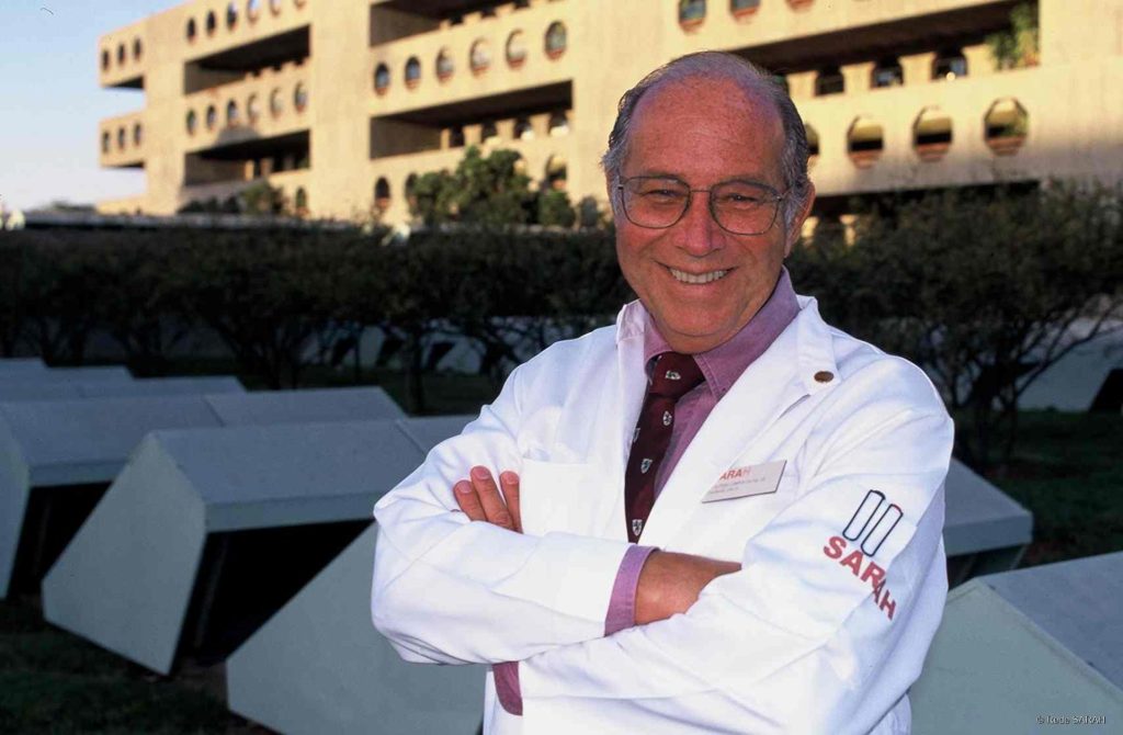 SARAH Brasília – 12/05/1997 Vestindo jaleco branco, camisa lilás e gravata vinho, Dr. Aloysio Campos da Paz posa, sorridente e de braços cruzados, em frente ao SARAH Brasília Centro.