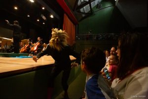 Integrante do Ballet de Brasília performando entre o público Em primeiro plano, no palco iluminado do Teatro SARAH, integrante do Ballet de Brasília dança em performance da fábula musical “Pedro e o Lobo”. Ao fundo, membros da Orquestra Sinfônica do Teatro Nacional Cláudio Santoro tocam seus instrumentos.