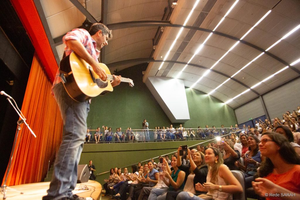Carlos Leoni tocando o violão À esquerda, de pé no palco do Teatro SARAH, Leoni toca violão e canta. À direita e ao fundo, plateia de pacientes, acompanhantes e colaboradores da Rede SARAH bate palmas, canta e tira fotos com celulares.