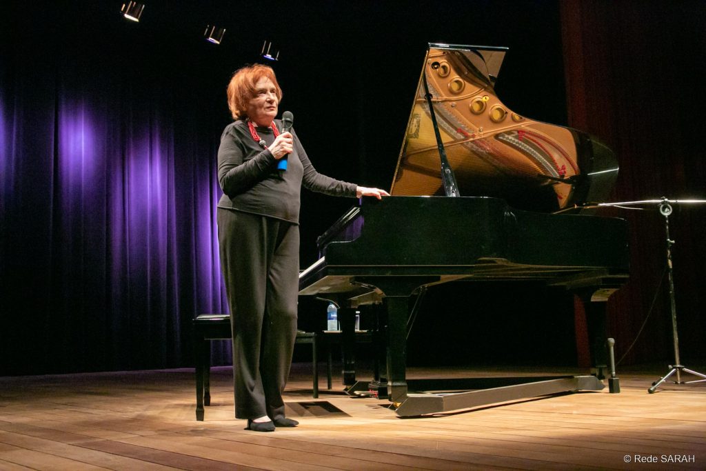 Clara Sverner conversando com o público De pé, no palco do Teatro SARAH, trajando roupas pretas e segurando microfone com a mão direita, a pianista Clara Sverner fala à plateia.