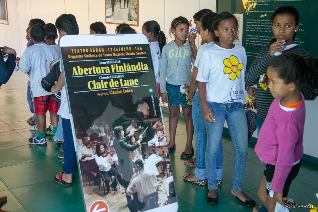 Crianças do Centro de Convivência Mozart Parada junto ao cartaz da apresentação Do lado de fora do Teatro SARAH, crianças do Centro de Convivência Mozart Parada porganizam-se em fila para assistir ao ensaio da Orquestra Sinfônica do Teatro Nacional Cláudio Santoro.