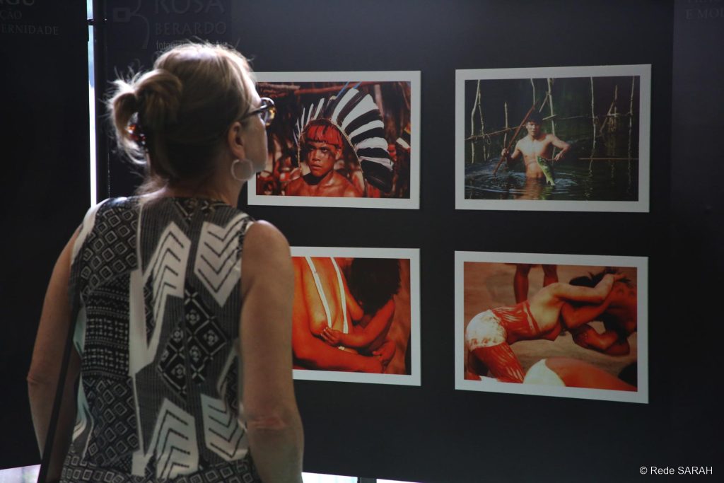 Convidada acompanhando a exposição De pé, mulher examina fotografias de indígenas brasileiros, coladas em um mural, como parte de exposição fotográfica no SARAH Brasília.