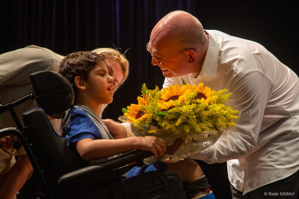 Luiz Blumenschein recebendo flores de um paciente do SARAH Brasília Sorrindo e trajando camisa social branca e calças pretas, o pianista Luiz Blumenschein recebe flores de menino em cadeira de roras, que traja uniforme de paciente hospitalar, no palco iluminado do Teatro SARAH Brasília.