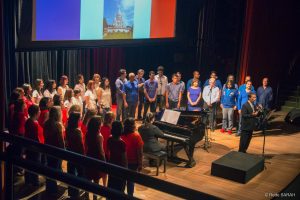 Maestro Carlos Ilha falando à plateia do Teatro SARAH No centro do palco, de pé e trajando terno escuro e gravata, o maestro Carlos Ilha fala à plateia do Teatro SARAH. Mais ao fundo, pianista sorri enquanto observa o maestro, sentada à frente de um piano de cauda. Ao seu redor, em um semicírculo, integrantes do Coral TST/Capes, todos de pé e trajando roupas vermelhas, azuis e brancas, em homenagem às cores da França, ouvem a fala do maestro atentamente.