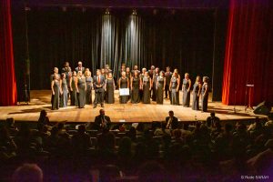 Coral do Senado performando Trajando ternos pretos com camisas brancas e gravatas vermelhas ou vestidos longos pretos, membros do Coral do Senado apresentam-se, de pé e dispostos em semicírculo no palco do Teatro SARAH.