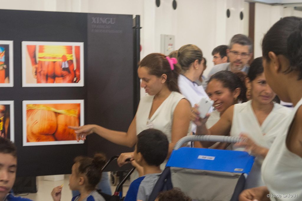 Pacientes, acompanhantes e colaboradores do SARAH São Luís acompanhando a exposição Vestindo uniforme da Rede SARAH, acompanhante aponta para a foto das nádegas de um indígena, exposta em um mural com os dizeres "Xingu", enquanto fala a crianças, também uniformizadas.