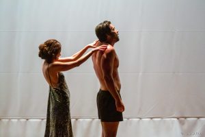 Sérgio Marone e Juliana Martins performando a peça “Eu te Amo” De pé, no palco do Teatro SARAH, em frente a um fundo branco, Juliana Martins, trajando um vestido longo de lantejoulas verdes, aparenta massagear as costas de Sérgio Marone, vestindo cueca tipo samba canção.