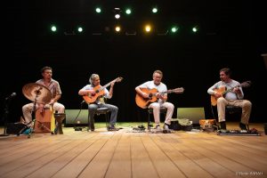 Da esquerda para direita Carlos Pial, Bosco Oliveira, Cacau Alencar e Manassés de Sousa Sentados lado a lado, no palco iluminado do Teatro SARAH apresentam-se três violonistas, trajando camisetas de algodão e calças compridas de diferentes cores e tecidos, e um percussionista, trajando camisa de mangas curtas com estampas florais e calças de tecido claro.