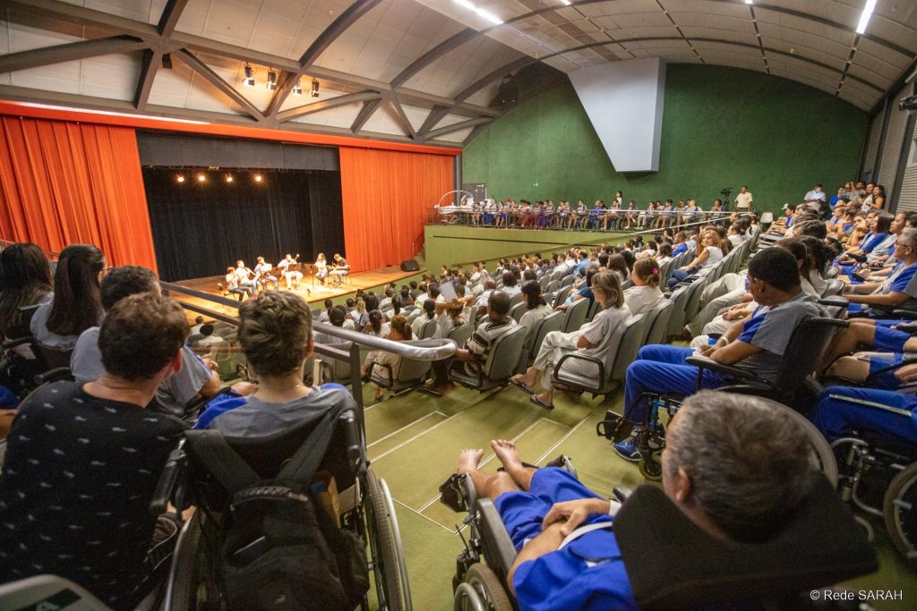 Público do Teatro SARAH Brasília assistindo à apresentação Em primeiro plano, nas galerias laterais e no auditório do Teatro SARAH, pacientes, acompanhantes e colaboradores da Rede, todos vestindo uniformes ou jalecos, acompanham a apresentação do Quinteto de Sopro e Percussão. Ao fundo, no centro do palco iluminado, os músicos tocam seus instrumentos.