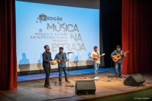 Grupo de Choro Sai da Frente performando juntos No palco iluminado do Teatro SARAH, de pé, apresentam-se quatro músicos, tocando pandeiro, cavaquinho, bandolim e violão. Trajam calças jeans, camisas de mangas compridas e tênis. Ao fundo, grande tela de projeção mostra os dizeres "8ª Edição – Música na Estrada, Música e Outras Artes Conectando as Pessoas”.
