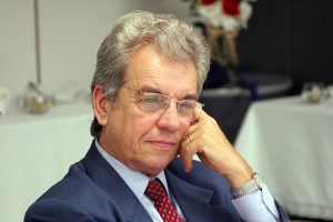 Ministro Carlos Átila Carlos Átila aparece sentado, vestindo terno azul-marinho e gravata vermelha, tocando o próprio rosto com a mão esquerda, em uma sala de reuniões.