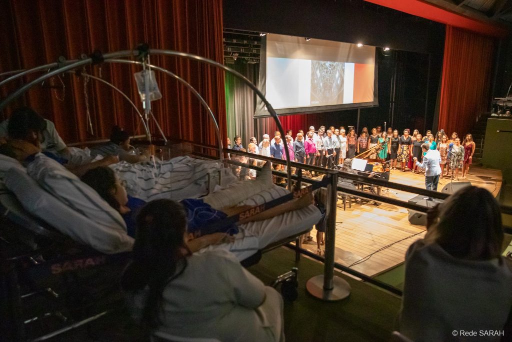 Pacientes do SARAH Brasília assistindo ao espetáculo Em primeiro plano, em uma das galerias laterais do Teatro SARAH, à sombra, três pacientes, deitados em macas e trajando uniformes hospitalares, acompanham o concerto “ITÁLIA – Grazie Mille”. Ao seu lado, sentadas em cadeiras, duas acompanhantes também assistem ao espetáculo. Ao fundo, no centro do palco iluminado, apresenta-se o Coral TST-Capes, regido pelo maestro Carlos Ilha.