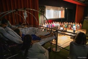 Pacientes do SARAH Brasília assistindo ao espetáculo Em primeiro plano, em uma das galerias laterais do Teatro SARAH, à sombra, três pacientes, deitados em macas e trajando uniformes hospitalares, acompanham o concerto “ITÁLIA – Grazie Mille”. Ao seu lado, sentadas em cadeiras, duas acompanhantes também assistem ao espetáculo. Ao fundo, no centro do palco iluminado, apresenta-se o Coral TST-Capes, regido pelo maestro Carlos Ilha.