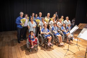 Ney Rosauro e Quarteto Capital posando para foto junto aos pacientes do SARAH Brasília Na coxia do Teatro SARAH, organizados em fileiras, Ney Rosauro e o Quarteto Capital posam para fotos com pacientes e acompanhantes. Em primeiro plano, cinco pacientes cadeirantes, trajando uniformes hospitalares. Logo atrás, cinco acompanhantes, também vestindo uniformes hospitalares. Ao fundo, Ney Rosauro e os cinco músicos do Quarteto Capital seguram ramalhetes de flores.