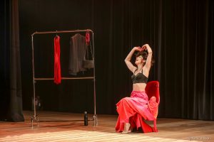 Juliana Martins apresentando a peça “O prazer é todo nosso” No centro do palco, sentada em uma cadeira vermelha, descalça e vestindo um top preto com uma saia vermelha longa e bufante, a atriz Juliana Martins sorri e levanta os braços, lançando ao ar os cabelos.