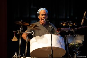 Sérgio Boré performando sua apresentação Close mostra o percussionista Sérgio Boré cantando ao microfone e tocando um tambor de aço. Iluminado, no palco do Teatro SARAH, ele traja camiseta cinza e boina azul-clara.
