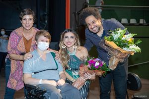 Atriz Maria Paula (à esquerda), Juliani Fernandes e Diego Figueiredo posando para foto junto à paciente do SARAH Brasília A atriz Maria Paula (à esquerda, vestindo calças jeans, uma bata estampada laranja e rosa, e uma blusa marrom), uma paciente do SARAH Brasília (sentada em cadeira de rodas, vestindo uniforme hospitalar), a cantora Juliani Fernandes (agachada, segurando um buquê de flores e trajando um vestido longo azul, verde e branco) e o violonista Diego Figueiredo (vestindo camisa de mangas longas azul e calças jeans) posam para foto no Teatro SARAH.
