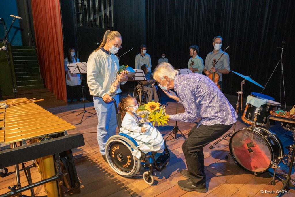 Ney Rosauro agradecendo as flores recebidas de uma paciente do SARAH Brasília Na coxia do Teatro SARAH, vestindo calças pretas e camisa de mangas compridas roxa com estampa floral, Ney Rosauro abaixa-se para receber flores de criança em cadeira de rodas, paciente do SARAH Brasília. Mais atrás, acompanhante da criança observa a cena. Ao fundo aparecem membros do Quarteto Capital, segurando seus instrumentos.