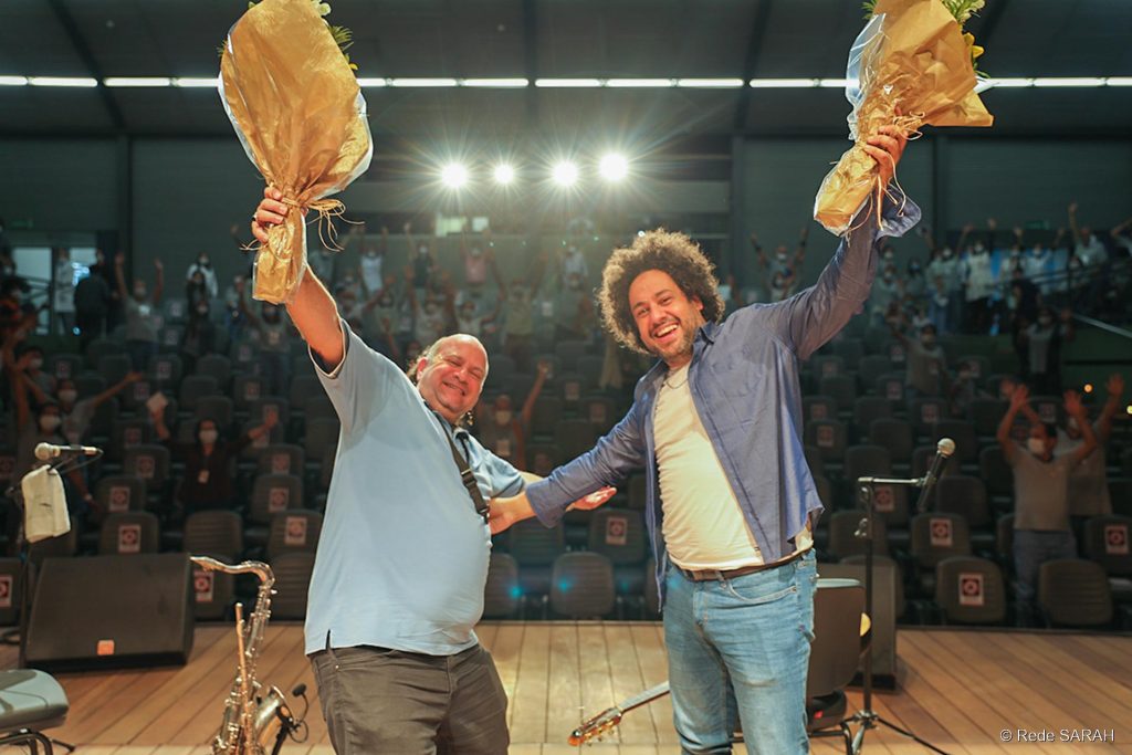 Derico Sciotti e Diego Figueiredo posando para foto junto ao público do Teatro SARAH De pé, sorrindo com os braços abertos e segurando buquês de flores, Diego Figueiredo e Derico posam no palco iluminado do Teatro SARAH em Brasília. Ao fundo, a plateia de colaboradores, pacientes e acompanhantes da Rede SARAH também ergue os braços e acena para a foto. Derico traja camisa polo azul-claro e calça de brim marrom; Diego veste calça jeans, camiseta branca e camisa social azul desabotoada.