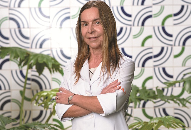 Dra Lúcia De pé e vestindo jaleco branco, Dra. Lúcia Willadino Braga posa para foto, de braços cruzados, em frente a azulejos de Athos Bulcão.