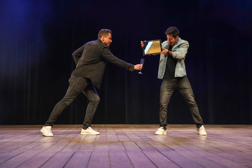 Ilusionistas Henry e Klauss performando juntos Ilusionistas Henry & Klauss apresentam-se no Teatro SARAH. Um deles segura uma taça vazia de espumante, enquanto o outro segura a tela de um tablet e parece derramar o conteúdo da tela na taça.