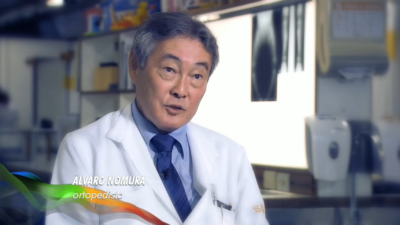 Matéria Superação – Caio Bonfim Dr. Álvaro Nomura aparece sentado e vestindo jaleco com camisa social azul e gravata azul-marinho. Ao fundo, consultório, onde aparecem radiografias de pacientes penduradas em tela luminosa.