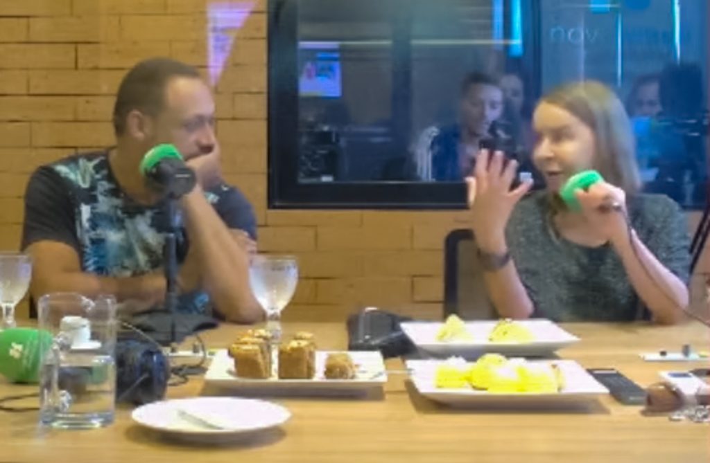 Dra. Lúcia Willadino Braga conversando com um dos apresentadores Captura de tela mostra imagem da transmissão, ao vivo, do programa de rádio Mesa pra Dois: sentada, à direita, Dra. Lúcia Willadino Braga conversa com membros da banda Moinho d’Água, todos sentados, em frente a microfones, ao redor de uma mesa com bebidas e lanche.