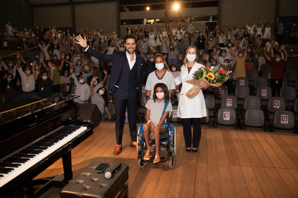 Thiago Arancam junto à Dra. Lúcia Willadino Braga e o público do Teatro SARAH posando para foto final do show Em fotografia tirada do fundo do palco do Teatro SARAH, o tenor Thiago Arancam, vestindo terno azul-marinho escuro e camisa social branca, posa com a presidente da Rede SARAH, vestindo jaleco branco, e uma paciente, em cadeira de rodas, e sua acompanhante. Ao fundo, pacientes, acompanhantes e colaboradores da Rede acenam da plateia para a foto.
