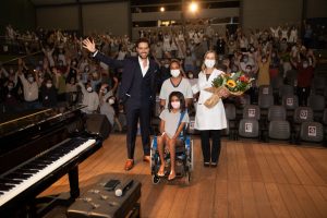Thiago Arancam junto à Dra. Lúcia Willadino Braga e o público do Teatro SARAH posando para foto final do show Em fotografia tirada do fundo do palco do Teatro SARAH, o tenor Thiago Arancam, vestindo terno azul-marinho escuro e camisa social branca, posa com a presidente da Rede SARAH, vestindo jaleco branco, e uma paciente, em cadeira de rodas, e sua acompanhante. Ao fundo, pacientes, acompanhantes e colaboradores da Rede acenam da plateia para a foto.