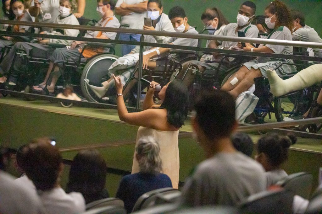 Vannick Belchior cantando para o público Vannick Belchior circula entre a plateia de pacientes, acompanhantes e colaboradores, enquanto canta, de pé e trajando um vestido branco longo, no Teatro SARAH.