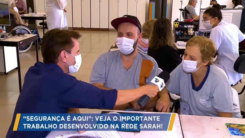 Maria Dalzotto, acompanhante de Zaqueu, paciente do SARAH Brasília, dando entrevista à Record TV Sentado e vestindo camisa azul-marinho, repórter segura microfone para ouvir acompanhante de paciente da Rede SARAH, que veste uniforme cinza. Entre eles, paciente, com uniforme cinza e boné preto, ouve atentamente. Todos usando máscaras cirúrgicas e sentados em torno de uma mesa. Ao fundo, pacientes e profissionais de reabilitação interagem.