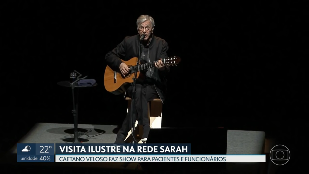 Matéria do DF2 da TV Globo Brasília Captura de tela da TV Globo Brasília mostra Caetano Veloso sentado, trajando casaco, camisa e calças pretas, cantando ao microfone e tocando violão, no centro do palco iluminado do Teatro SARAH. Na base da imagem, vinheta mostra os dizeres: "VISITA ILUSTRE NA REDE SARAH. Caetano Veloso faz show para pacientes e funcionários."