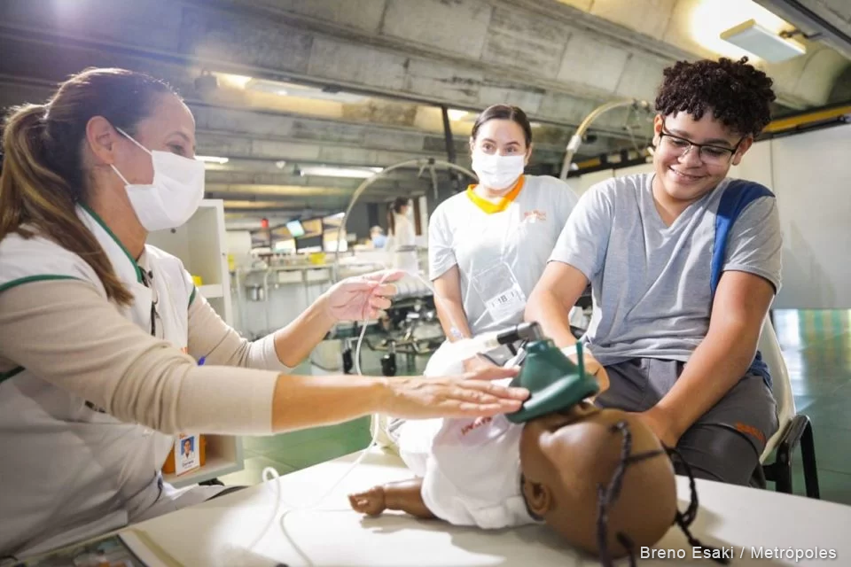 Profissional de saúde do SARAH Brasília utilizando a boneca para explicar o procedimento cirúrgico a paciente infantil (Crédito: Bruno Esaki, Metrópoles) Foto tirada em enfermaria do SARAH Brasília. Sentada, profissional de saúde, usando máscara cirúrgica e vestindo uniforme, manuseia boneca de plástico, que tem um bocal de anestesia verde no rosto. Ao seu lado, menino com uniforme de paciente sorri e examina a boneca. Mais atrás, mulher com uniforme de acompanhante e máscara cirúrgica observa a cena. (Crédito: Bruno Esaki, Metrópoles).